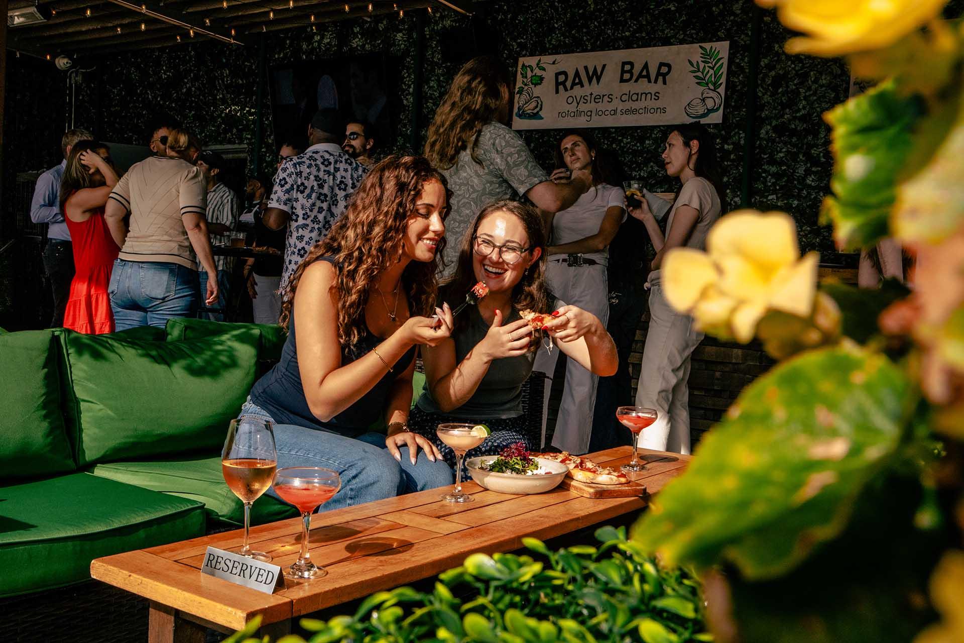 Two women sit on a green couch, smiling and eating seafood at an outdoor raw bar. Drinks and food are on the wooden table labeled “reserved.” People and plants surround the lively scene.