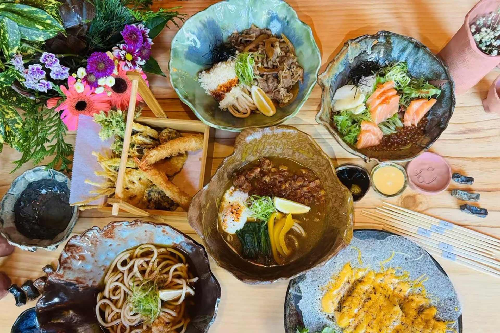A variety of Japanese dishes, including udon, curry rice, sashimi, fried tempura, and katsu, are arranged on a wooden table with a bouquet of flowers and chopsticks beside the plates.