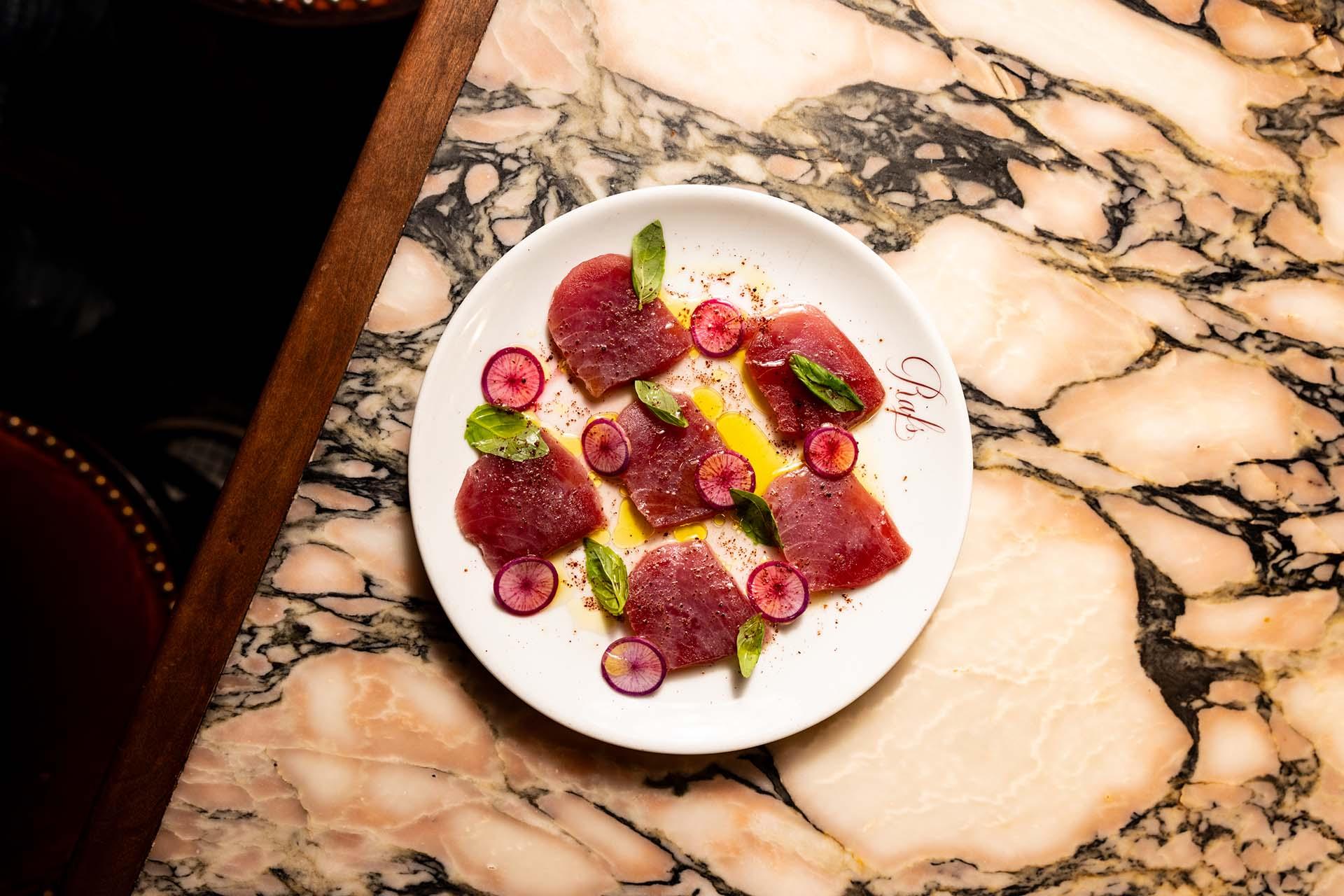 A white plate with slices of raw tuna, sliced radishes, basil leaves, and a few yellow strips, set on a marble tabletop.