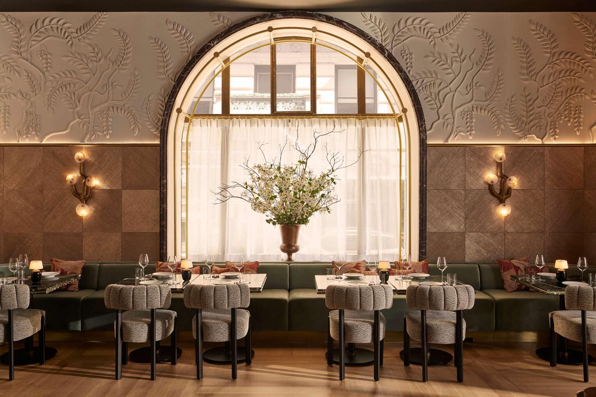 Elegant restaurant interior with a large arched window, textured walls, a long green banquette, neatly set tables, soft lighting, and a tall floral arrangement in the center.