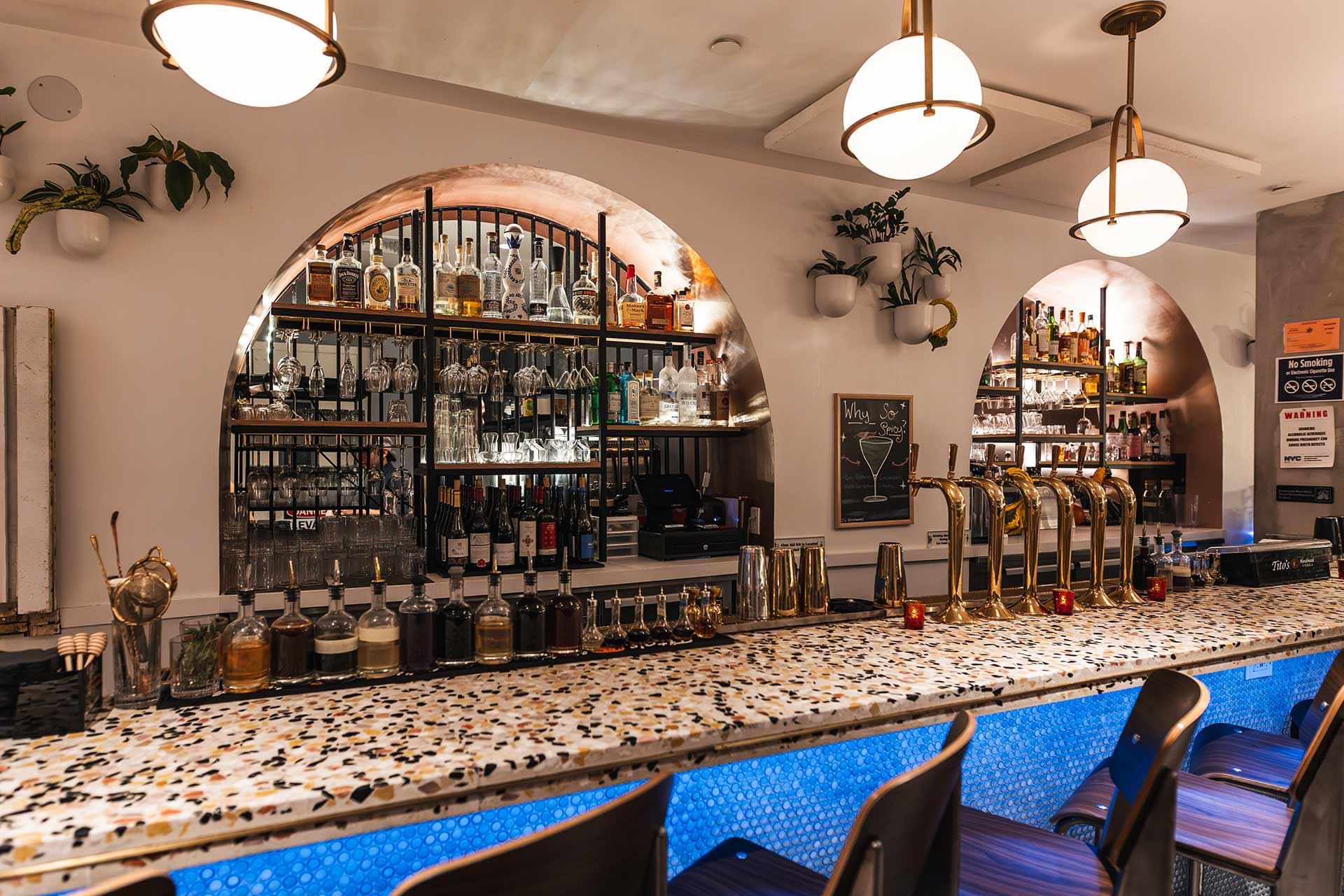 A stylish bar with a terrazzo countertop, blue accent lighting, gold beer taps, and shelves filled with bottles of alcohol against arched mirrors. Modern pendant lights and potted plants decorate the space.