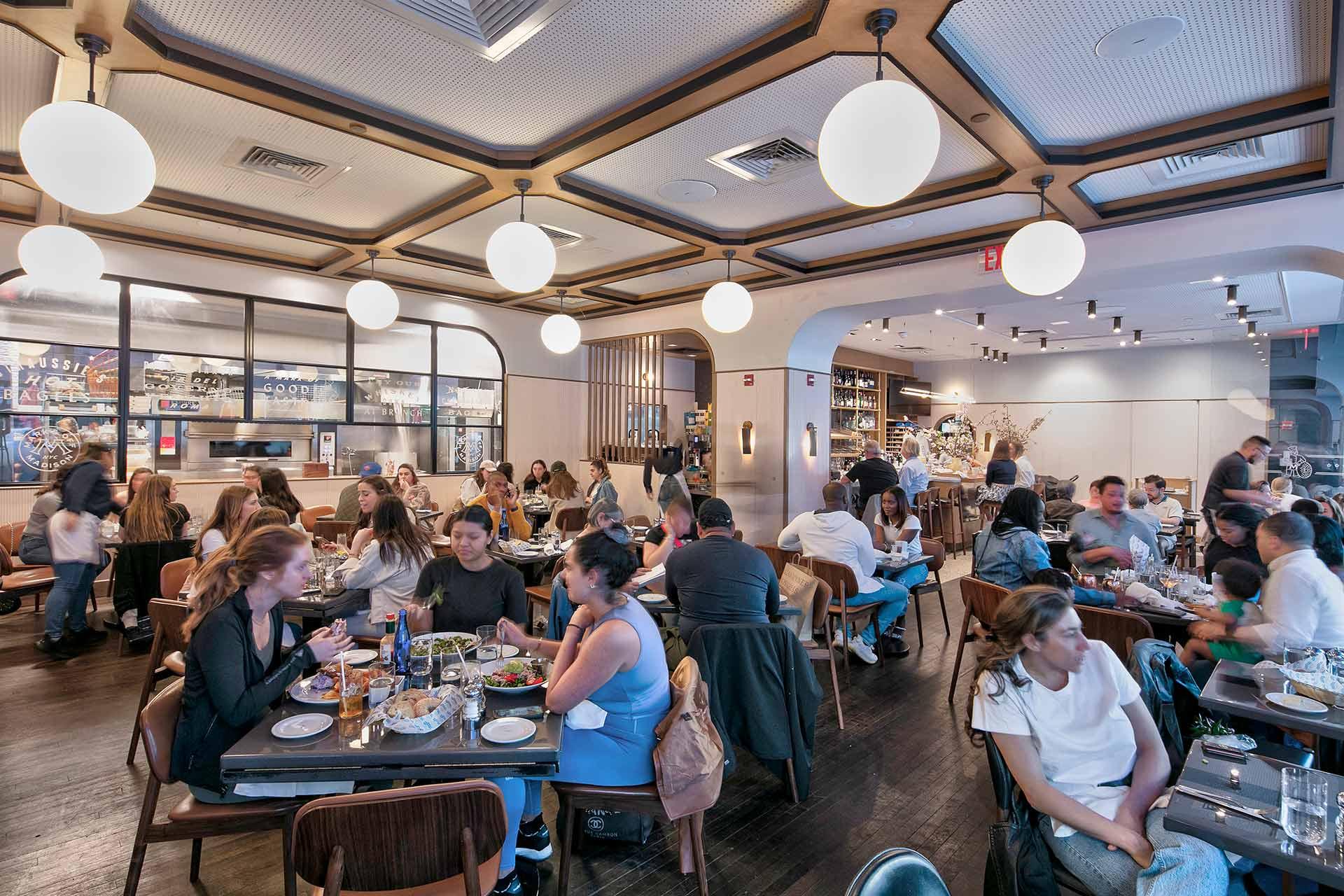 A busy, modern restaurant with people dining at tables, enjoying meals and conversation under round hanging lights. The atmosphere is lively, with staff serving food and a view of the open kitchen in the background.