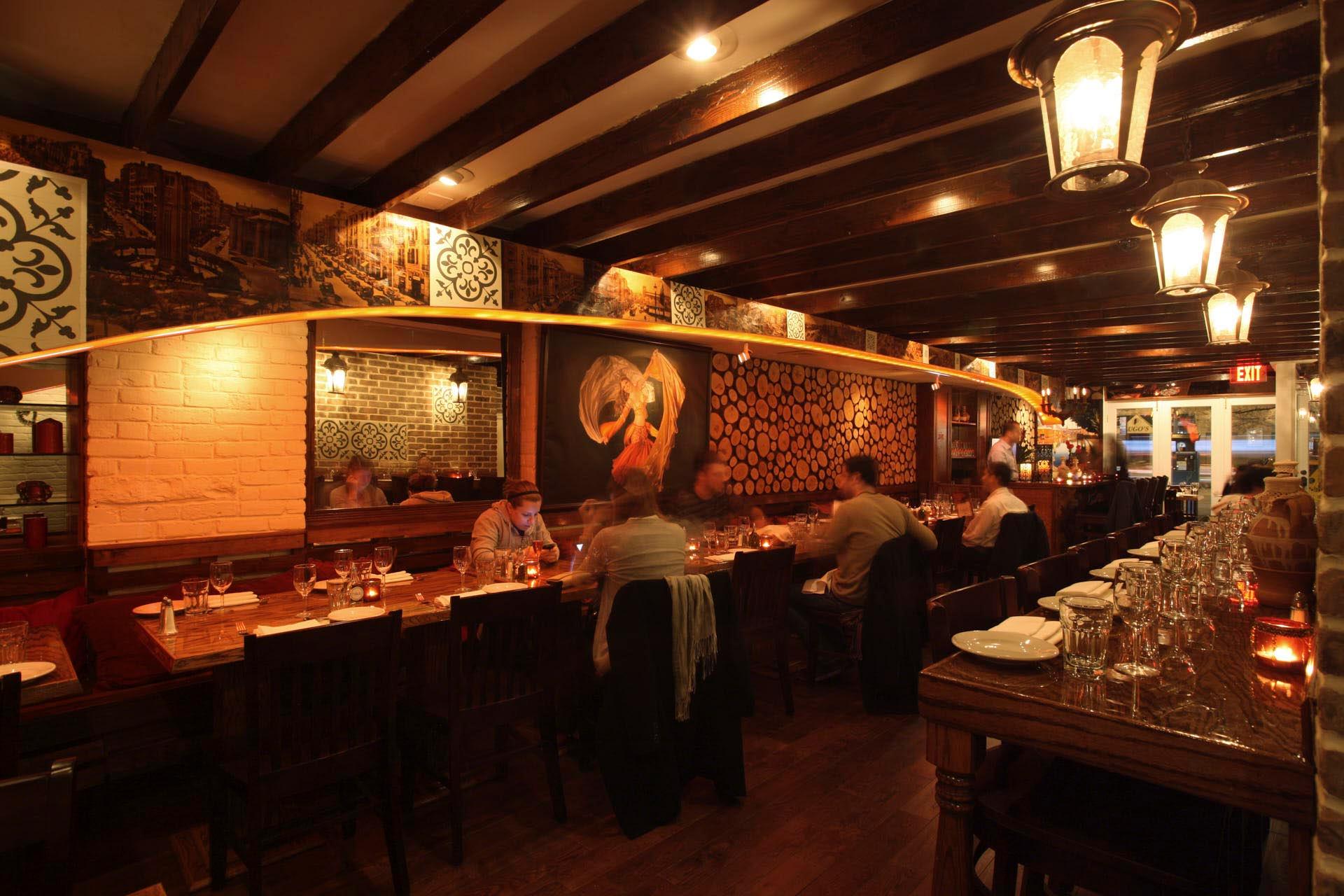 A warmly lit restaurant interior with wooden beams on the ceiling, people dining at tables along a brick wall adorned with paintings and decorative patterns, and glasses set on the tables.