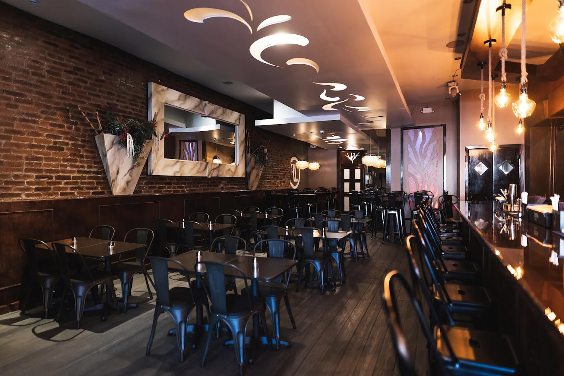 A modern restaurant interior with exposed brick walls, dark wooden furniture, decorative mirrors, and artistic light fixtures. Rows of empty black chairs and tables are neatly arranged, and pendant lights hang above the bar area.