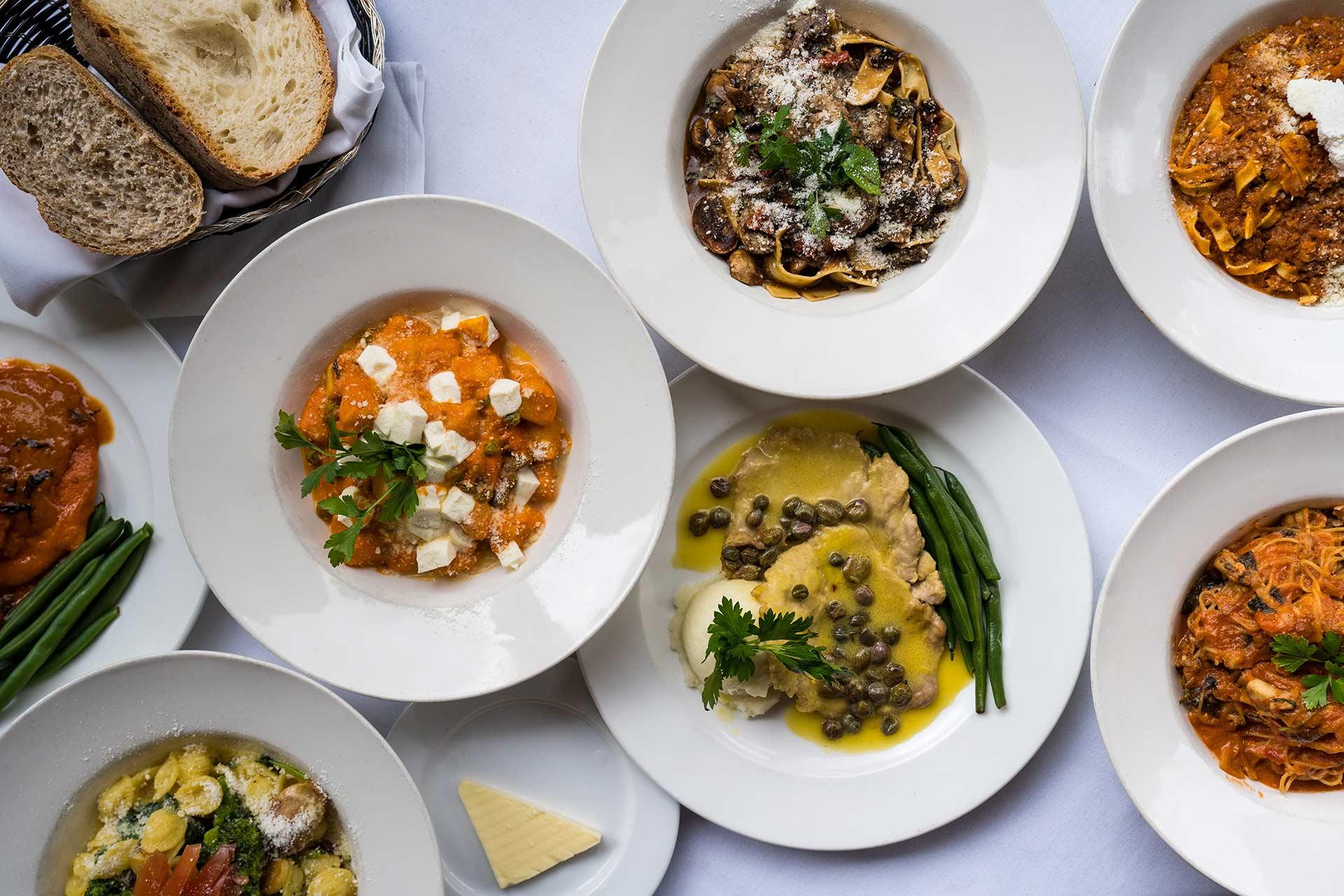 An overhead view of plates with various Italian dishes, including pasta with red and white sauces, ravioli with green beans, gnocchi, bread slices, and a serving of butter, all arranged on a white tablecloth.