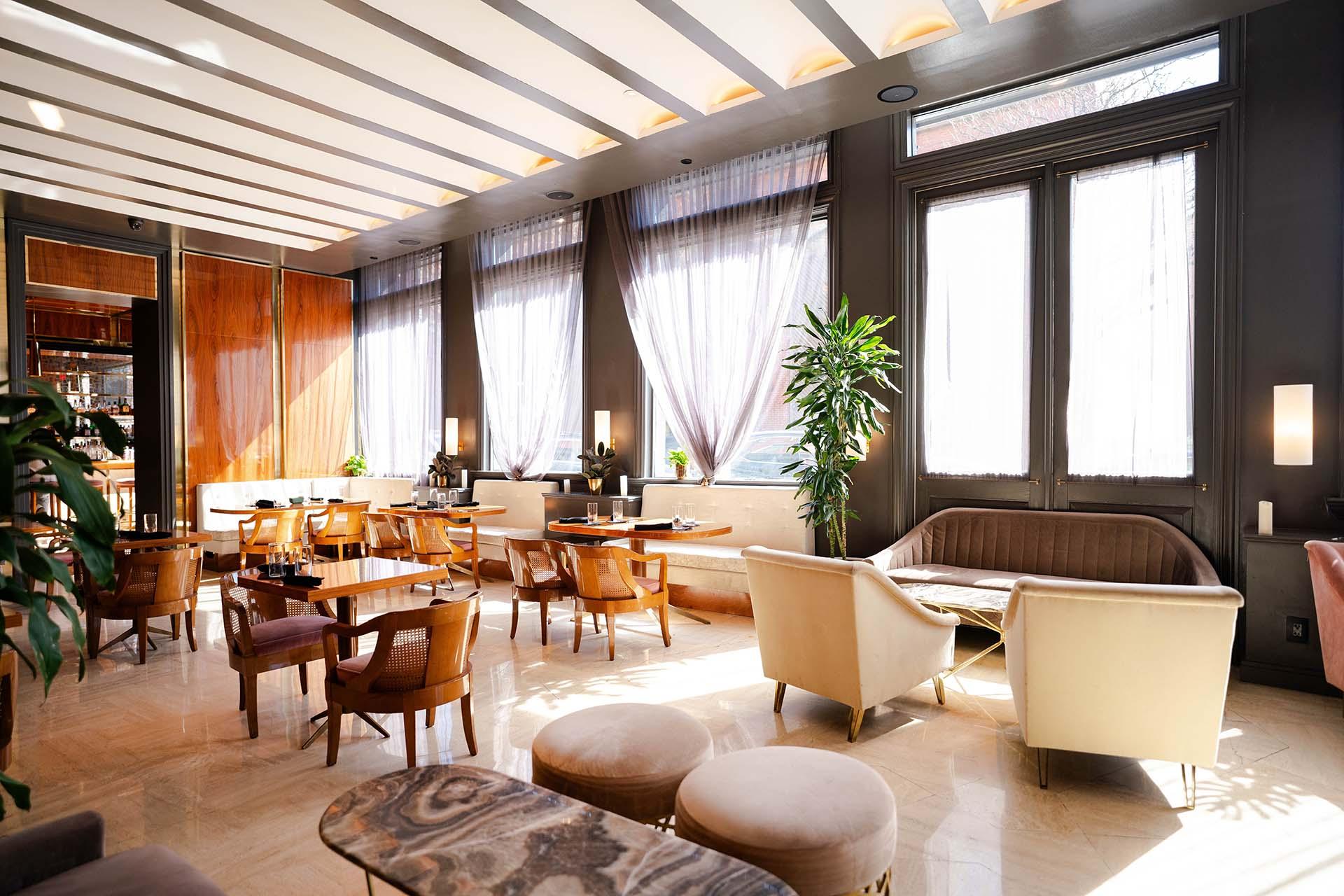 Bright, modern lounge with large windows, sheer curtains, stylish chairs, sofas, and tables. Light floor, dark walls, indoor plants, and soft lighting create a welcoming atmosphere.