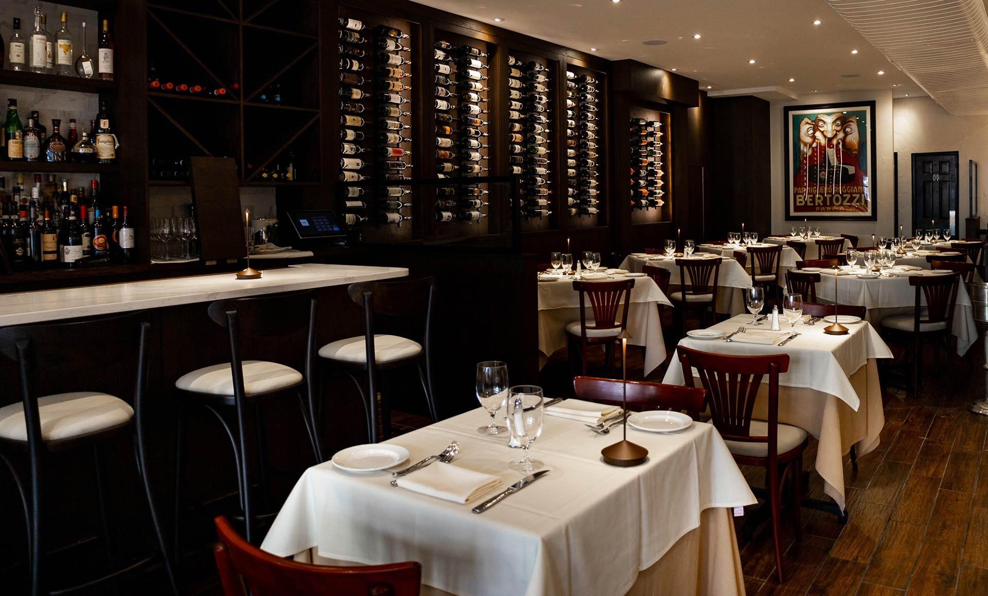 Elegant restaurant interior with white tablecloths, wooden chairs, wine racks on the wall, a stocked bar, and a framed poster. The space is softly lit and neatly arranged for dining.