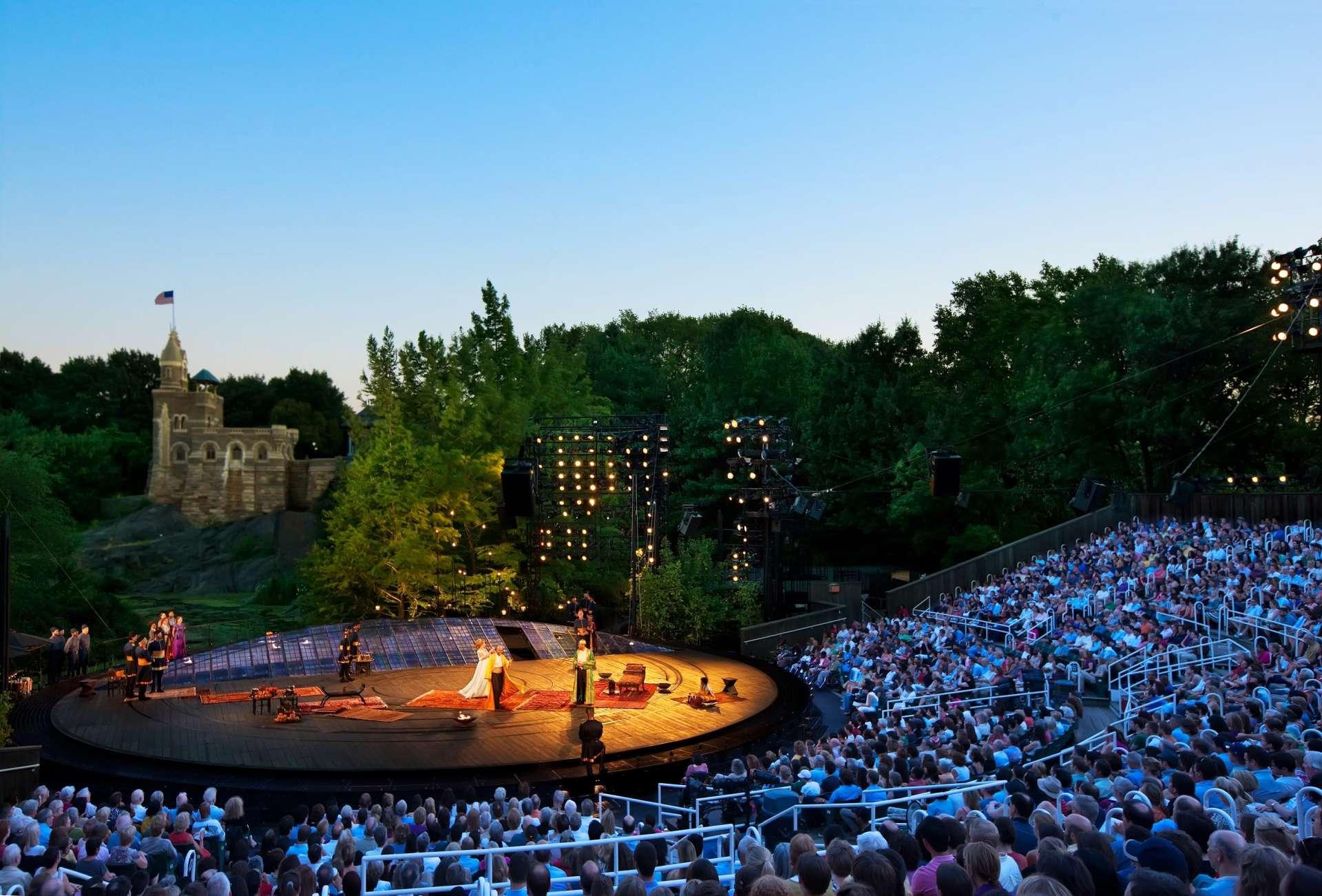 A large outdoor theater packed with people faces a round stage, where performers stand under bright lights. Trees and a castle-like building are visible in the background against a clear evening sky.