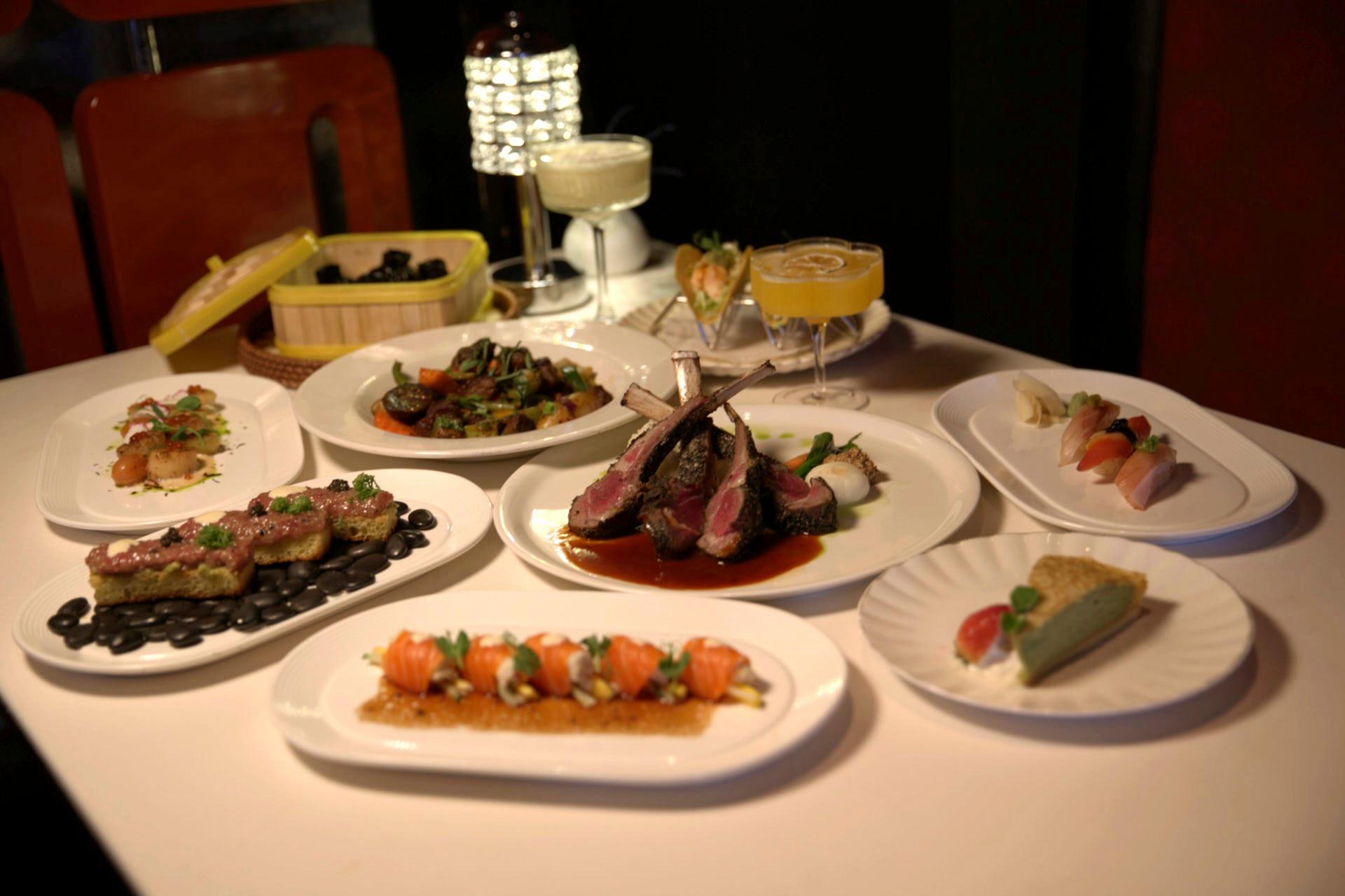 A table with a variety of gourmet dishes, including sliced meat, sushi, desserts, cocktails, and vegetables. Plates are arranged neatly with artistic presentations, set in a dimly lit elegant dining environment.