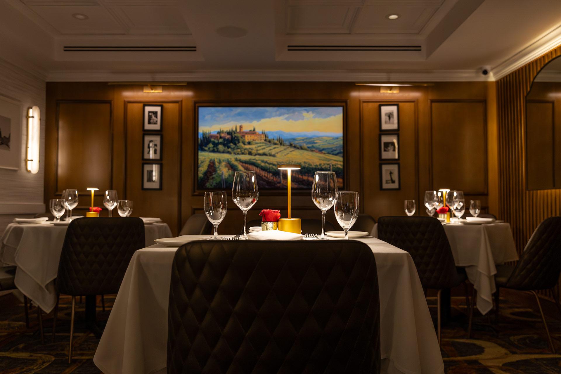 A dimly lit, elegant restaurant with tables set with white cloths, wine glasses, and a single red rose centerpiece. A large landscape painting and framed photos decorate the brown-paneled walls.