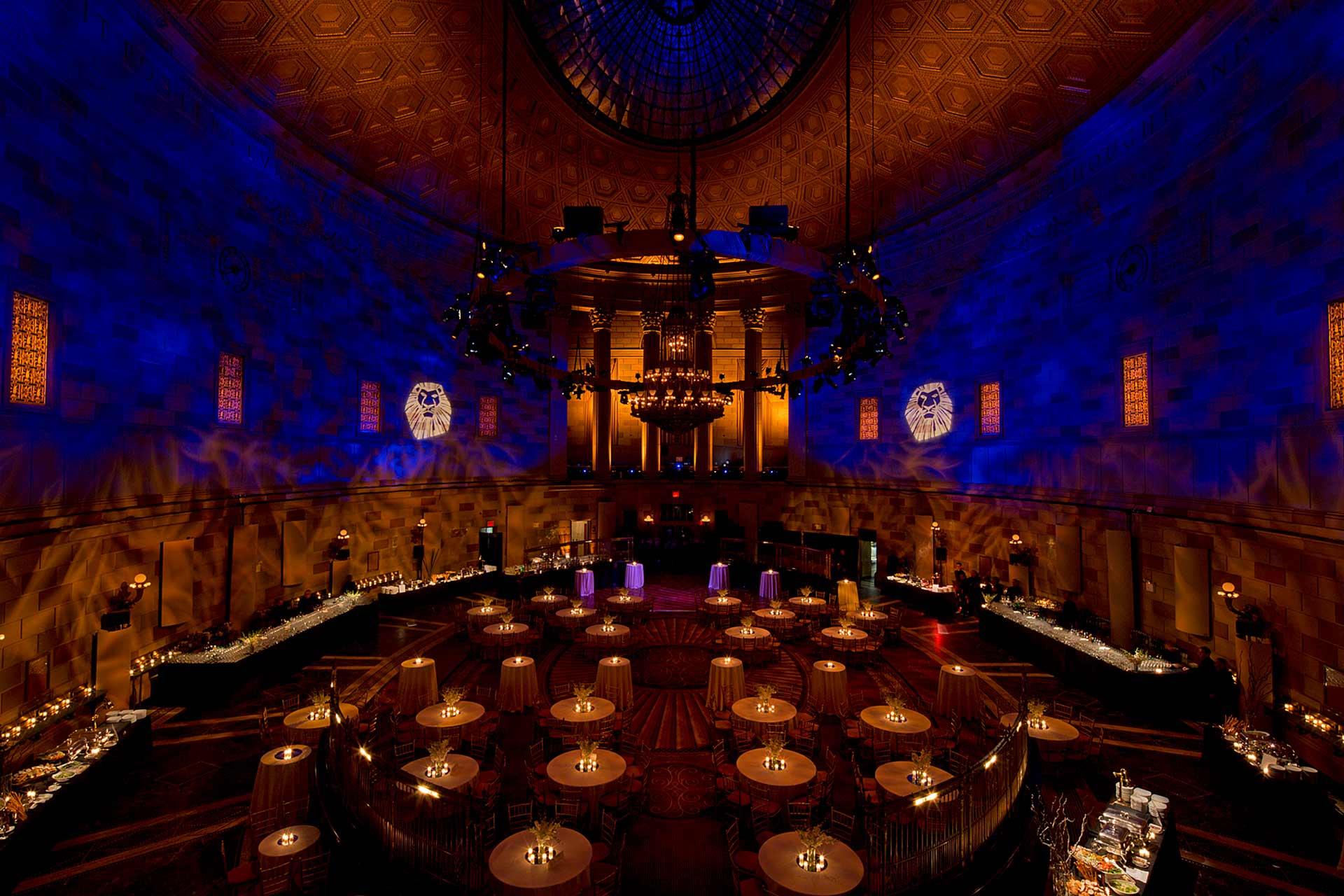 Grand ballroom with a domed ceiling lit by dramatic blue and gold lighting, round tables arranged below, and elegant decor along the walls, set for a formal event or banquet.