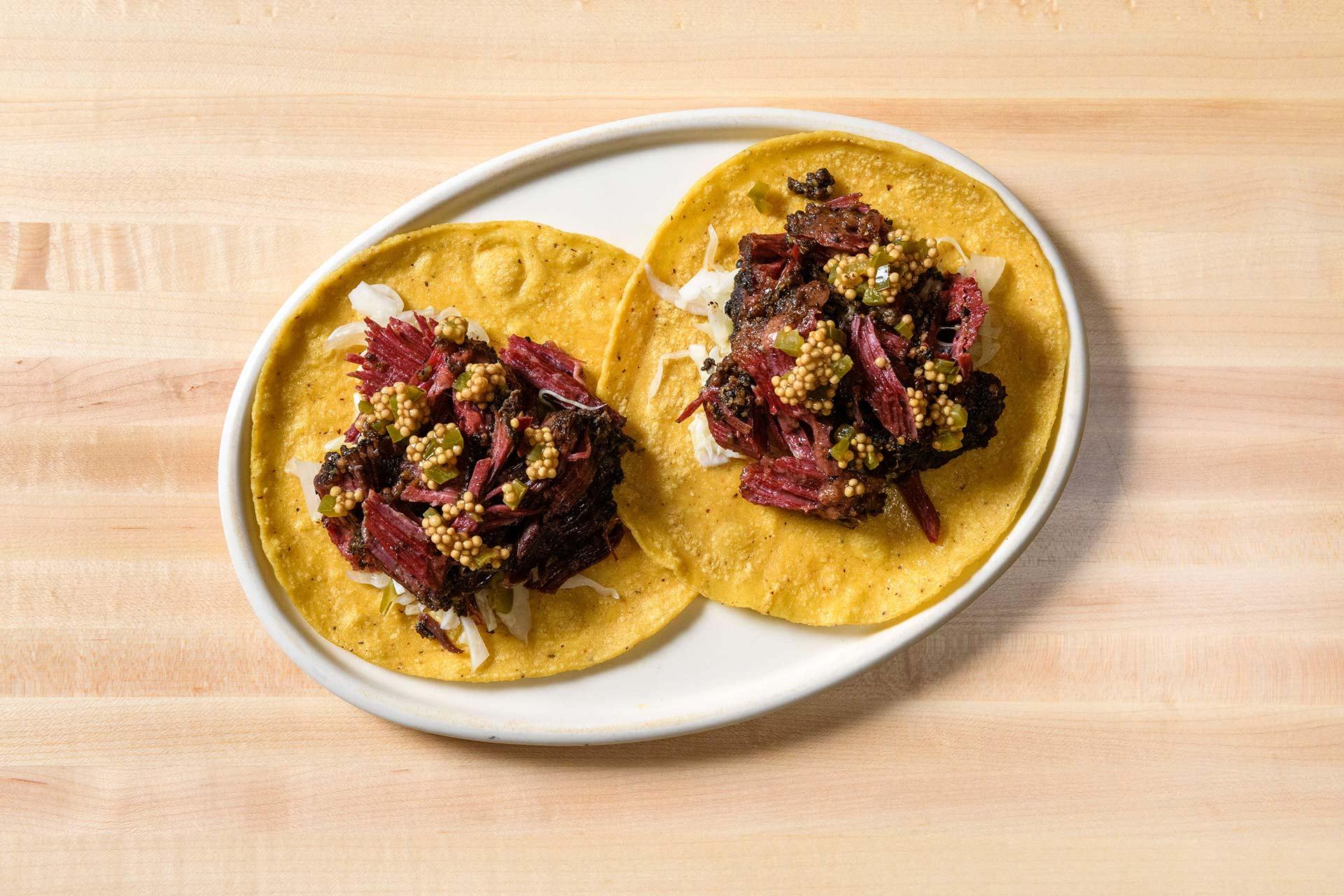 Two yellow corn tortillas topped with shredded meat, chopped onions, and pickled mustard seeds on an oval white plate, placed on a light wooden surface.