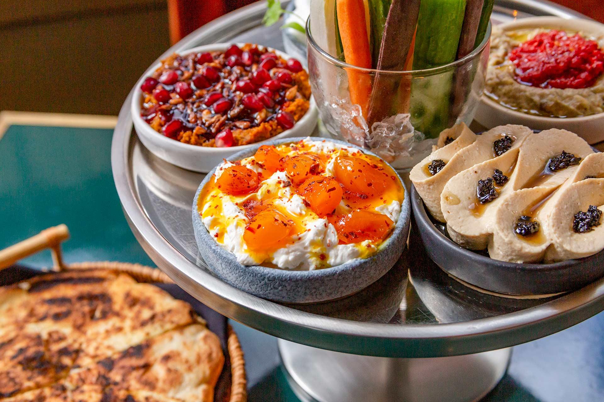A tiered tray with assorted Middle Eastern appetizers, including labneh with roasted tomatoes, hummus, baba ghanoush with red pepper, a bean salad, sliced bread, and a glass of fresh vegetable sticks.