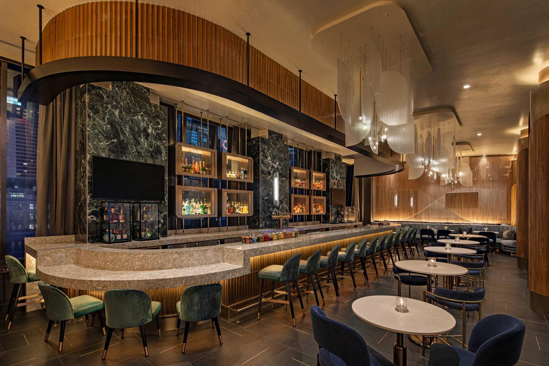 Sleek modern bar interior with a marble counter and high stools on the left, and tables with chairs on the right. The space features dim lighting, wooden accents, hanging light fixtures, and a wall-mounted display of bottles.