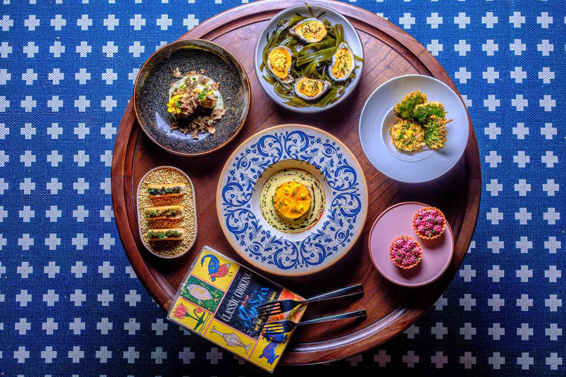 A round wooden table with six colorful plates of assorted gourmet appetizers, two spoons, and a vibrant illustrated menu, on a blue patterned tablecloth.