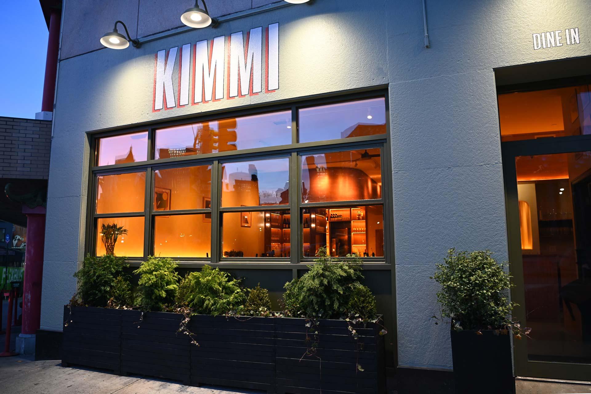 Restaurant exterior at dusk with large windows showing a warmly lit interior. "KIMMI" is displayed above the windows. Green plants in boxes line the front. "DINE IN" text is visible in the upper right corner.