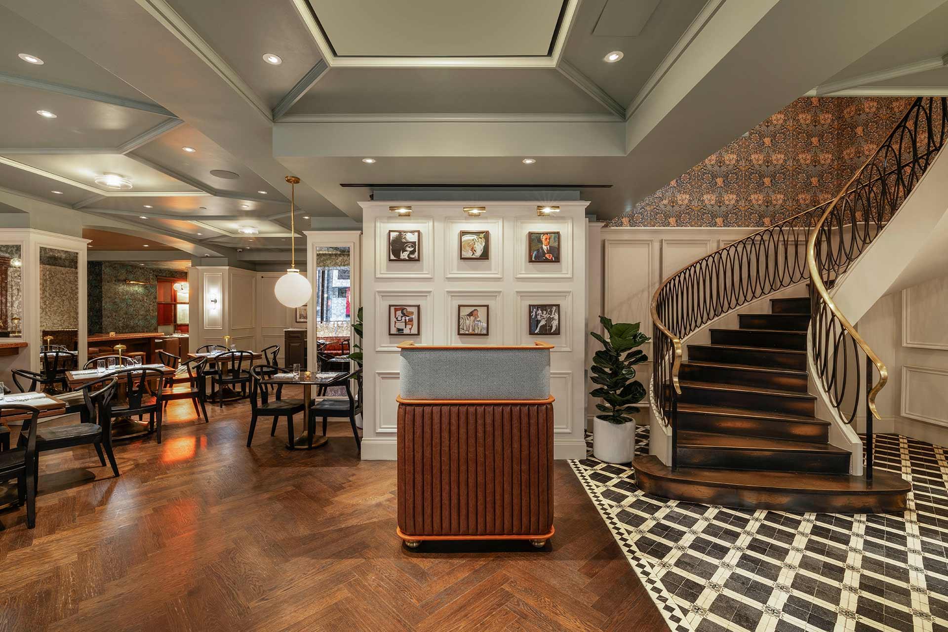 Elegant restaurant interior with a wooden host stand, framed photos on the wall, stylish wood staircase with a gold railing, patterned tile and wood flooring, and modern dining tables and chairs in the background.