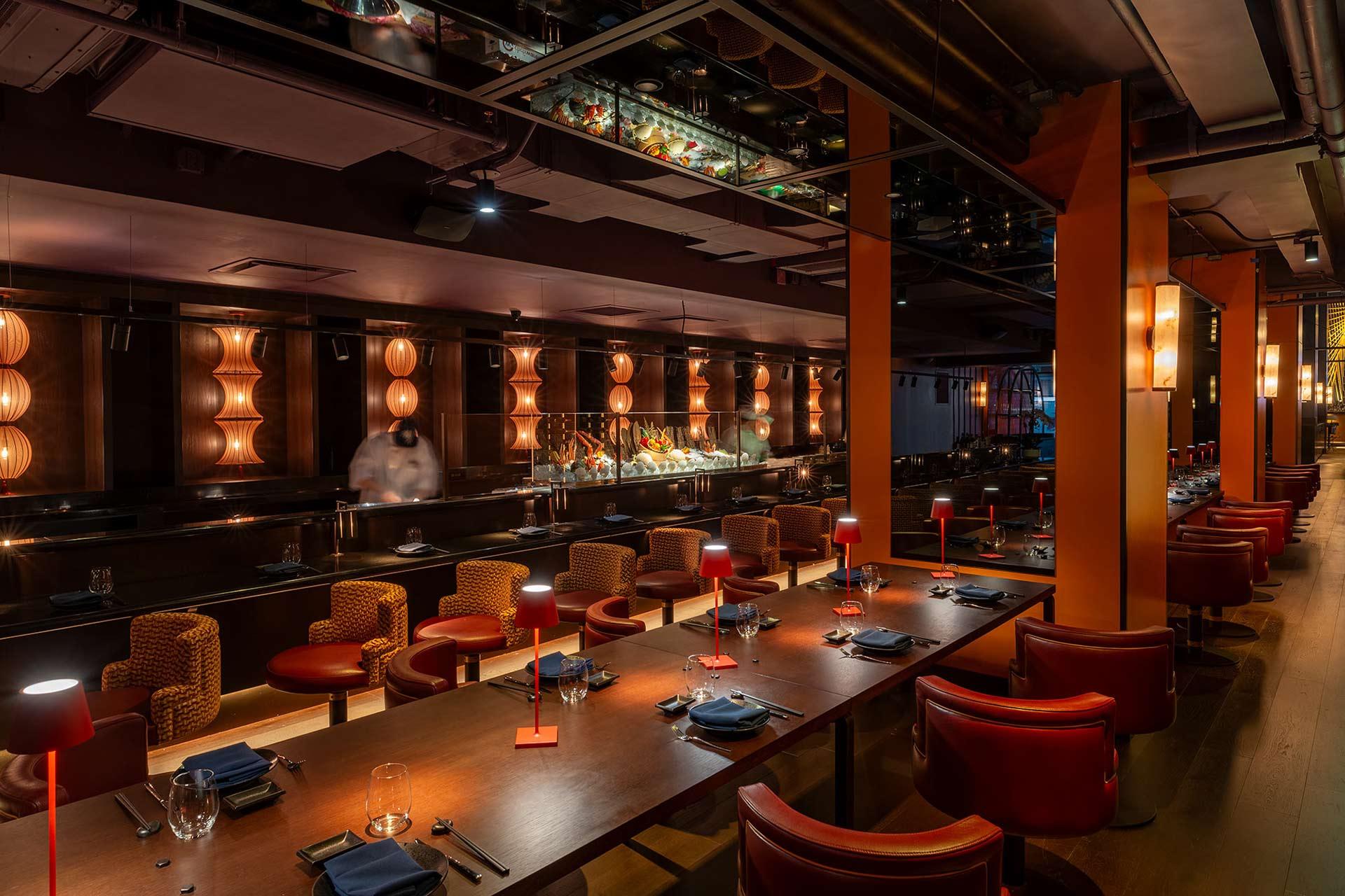 A stylish, dimly lit restaurant interior with long wooden tables, red leather chairs, and warm ambient lighting from modern wall fixtures and table lamps. The atmosphere is cozy and sophisticated.