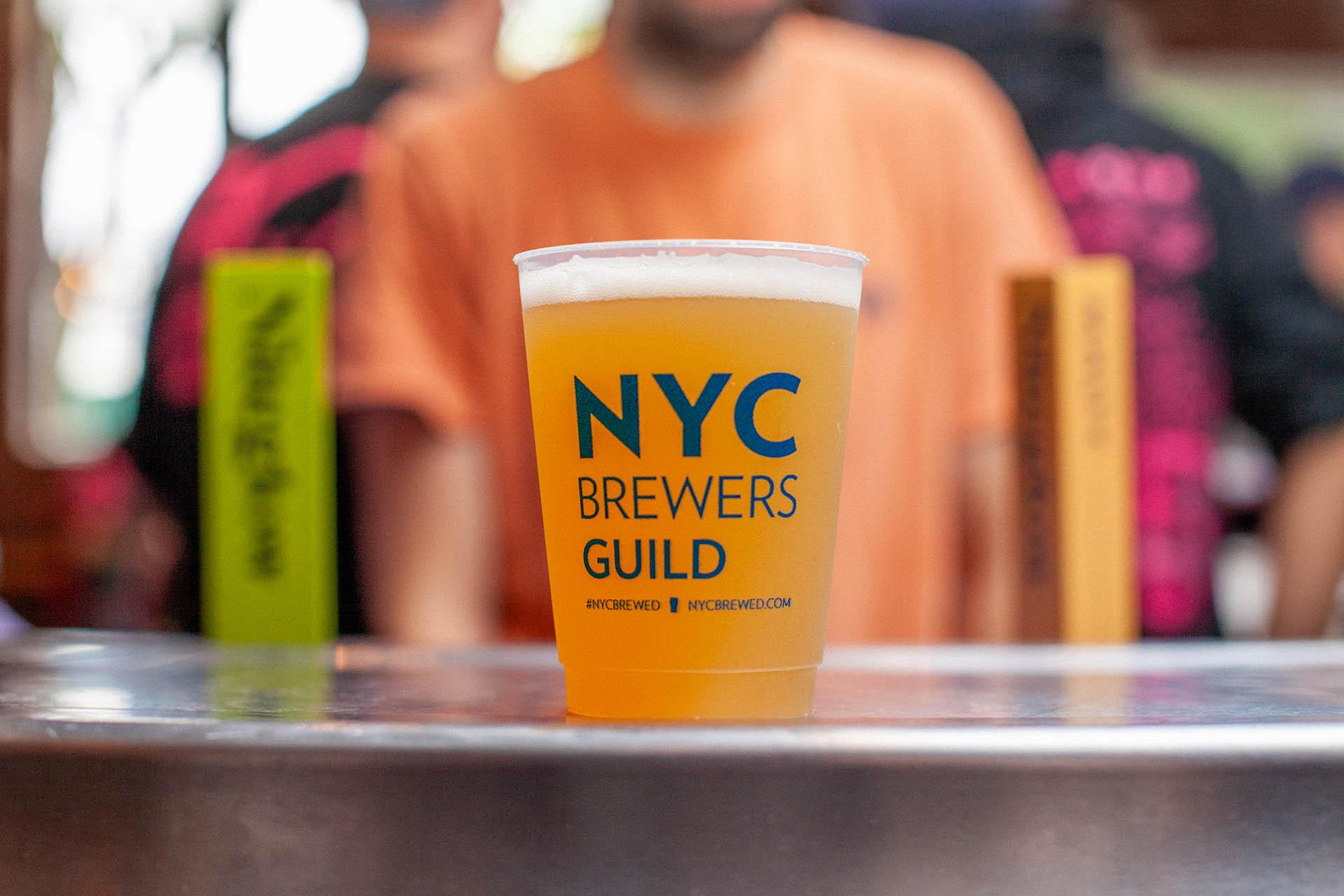 A plastic cup filled with beer, labeled “NYC Brewers Guild,” sits on a metal surface. The background shows people wearing colorful shirts, out of focus, and upright rectangular objects in green and orange.