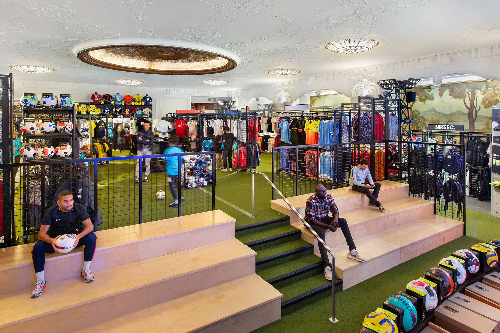 A spacious sports store interior with turf flooring, racks of colorful sports clothing, and shelves of soccer balls. Four people are sitting on wooden bleachers, and displays of athletic gear fill the brightly lit room.