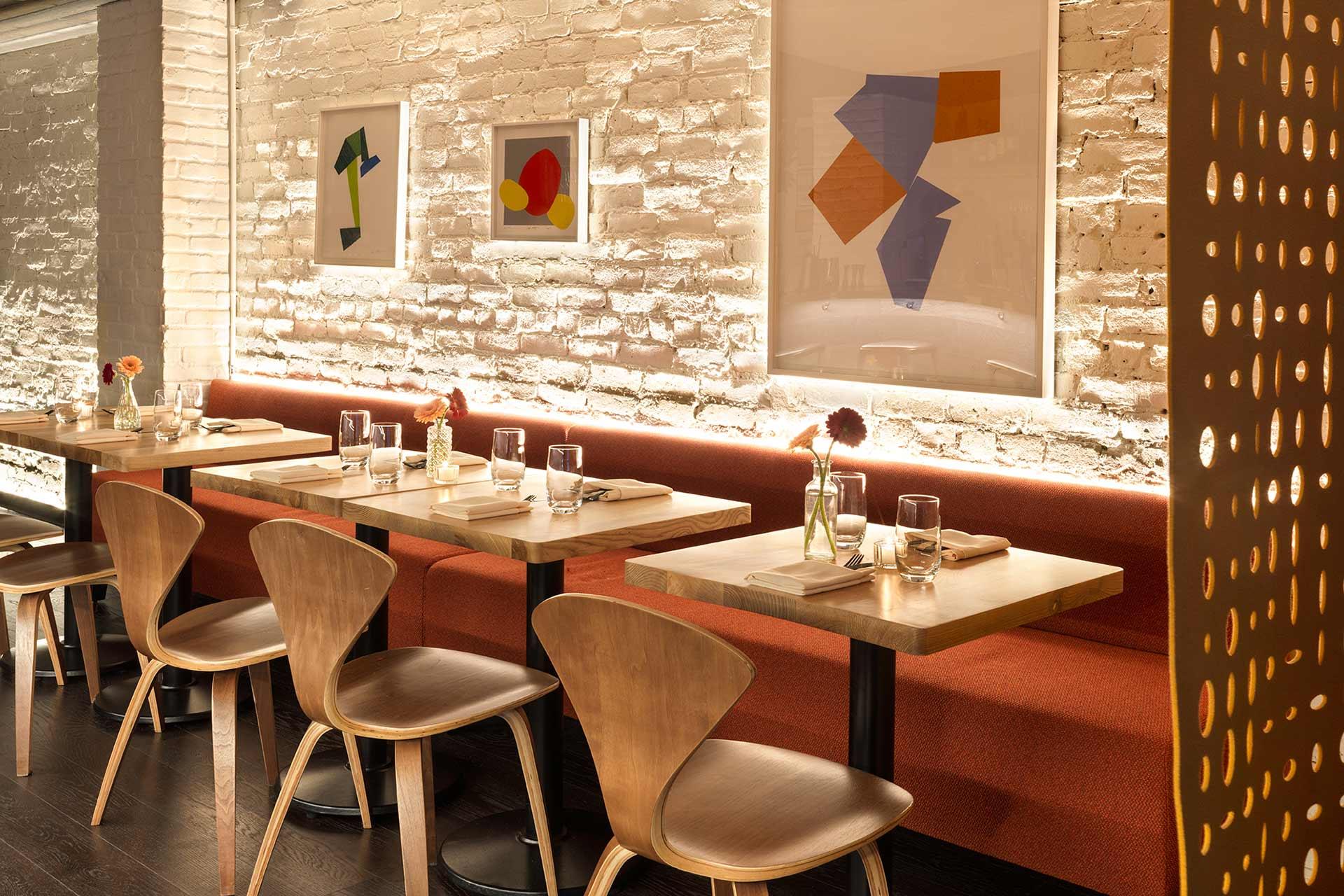 A cozy restaurant interior with wooden tables and chairs, red cushioned bench seating, exposed white brick walls, modern abstract art, and small vases with flowers on each table, softly lit by warm lighting.