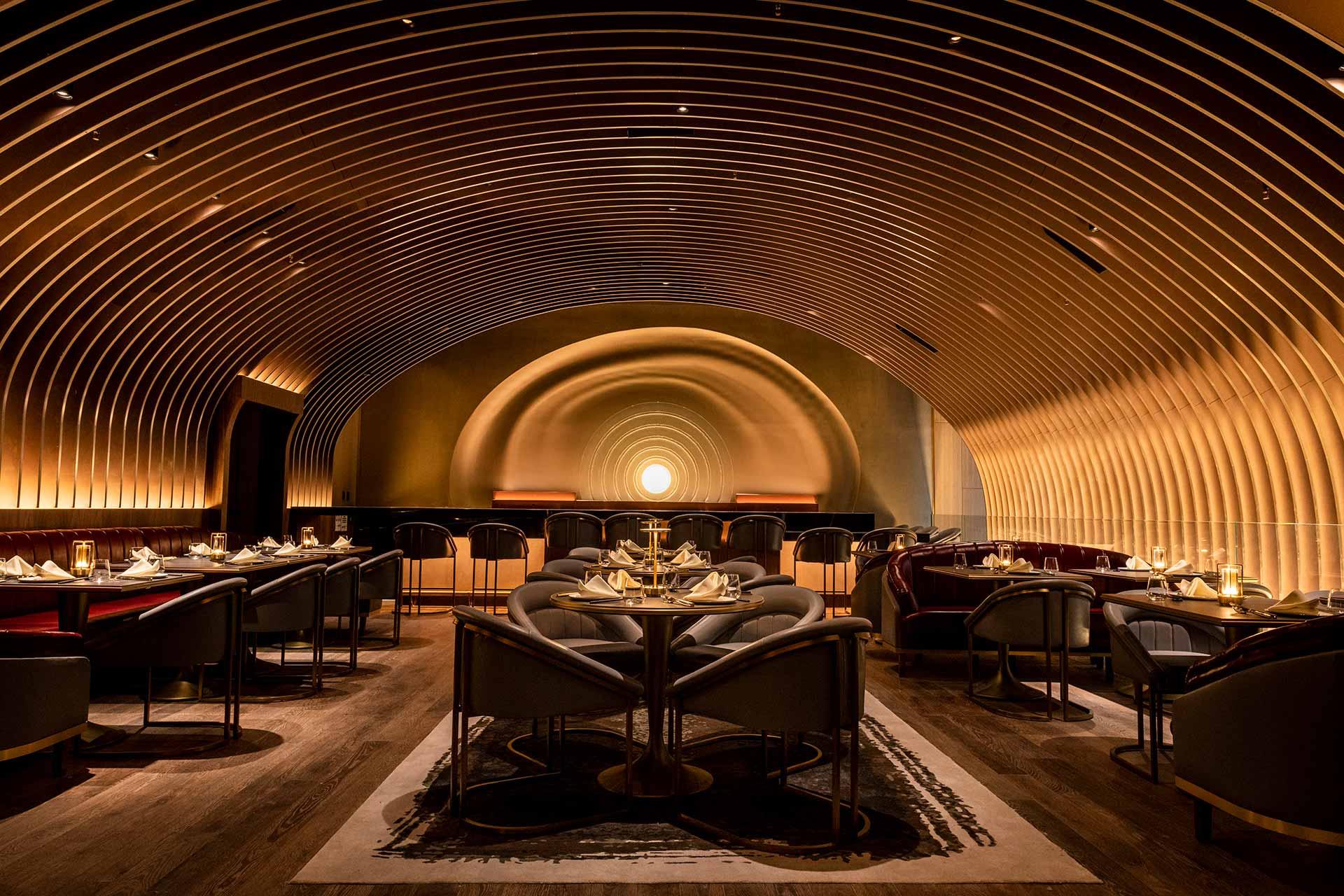 A luxurious restaurant interior with curved, tunnel-like golden arches, elegant lighting, round tables set for dining, and plush gray chairs on a wooden floor. The ambiance is warm and sophisticated.