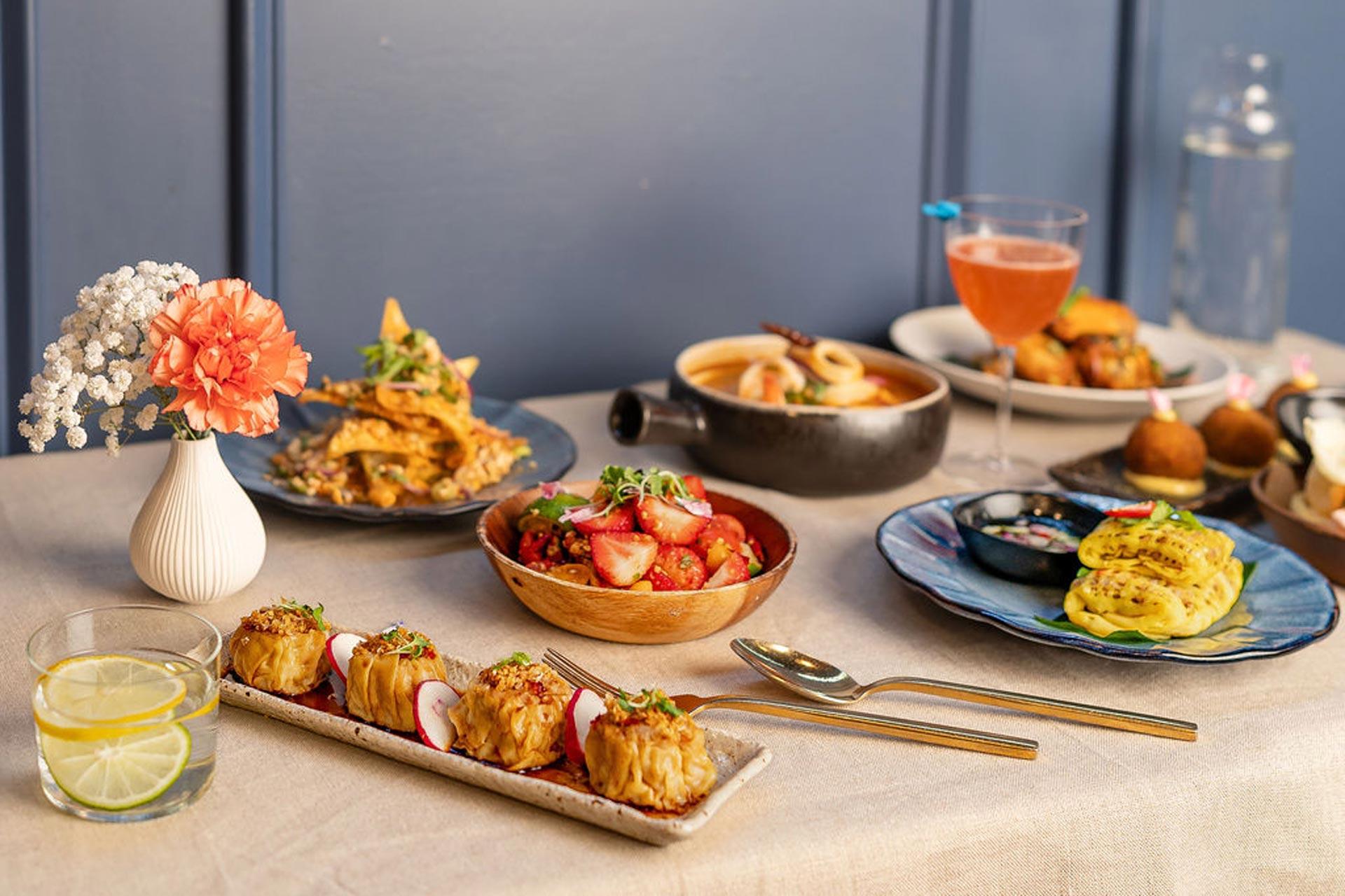 Alt Text A table set with an assortment of dishes including dumplings, a fruit salad, a casserole, and small burgers. A glass of lemon water and a pink cocktail accompany the meal, with a small floral arrangement nearby.