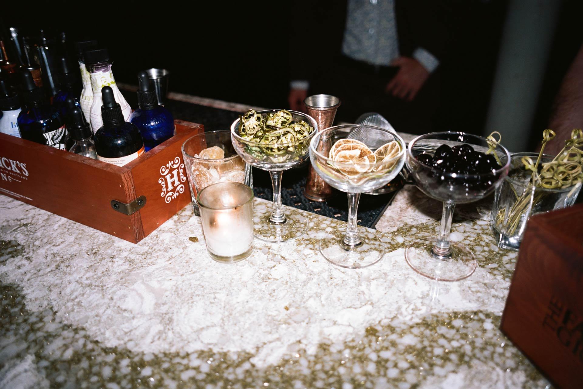 A bar counter with bottles, cocktail garnishes in coupe glasses, a candle, and a box of tools. A person in a suit is partially visible behind the counter.