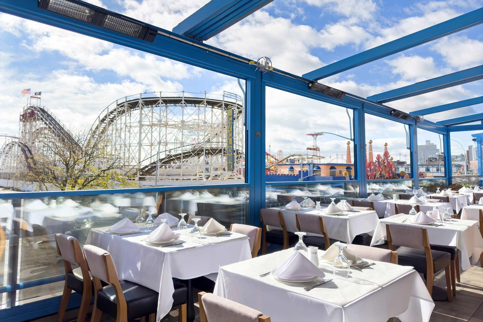A bright outdoor restaurant with neatly set tables, white tablecloths, and views of a wooden roller coaster and amusement park under a blue sky with scattered clouds.
