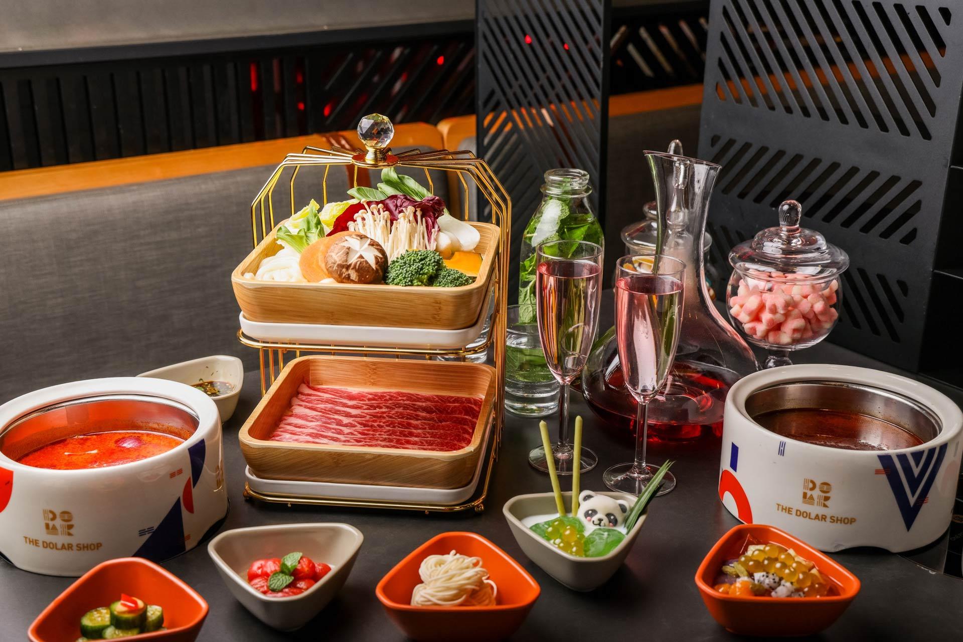 A table set for hot pot with two bowls of broth. Ingredients include sliced meat, mushrooms, and vegetables on a tiered tray. Various dipping sauces and small bowls of condiments are arranged around the table. Two glasses of rosé are also present.