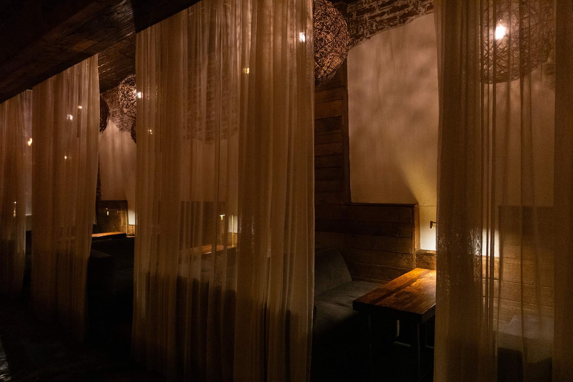 Dimly lit restaurant booths separated by sheer curtains, with wooden tables, cushioned benches, soft warm lighting, and rustic wooden and brick walls creating an intimate, cozy ambiance.