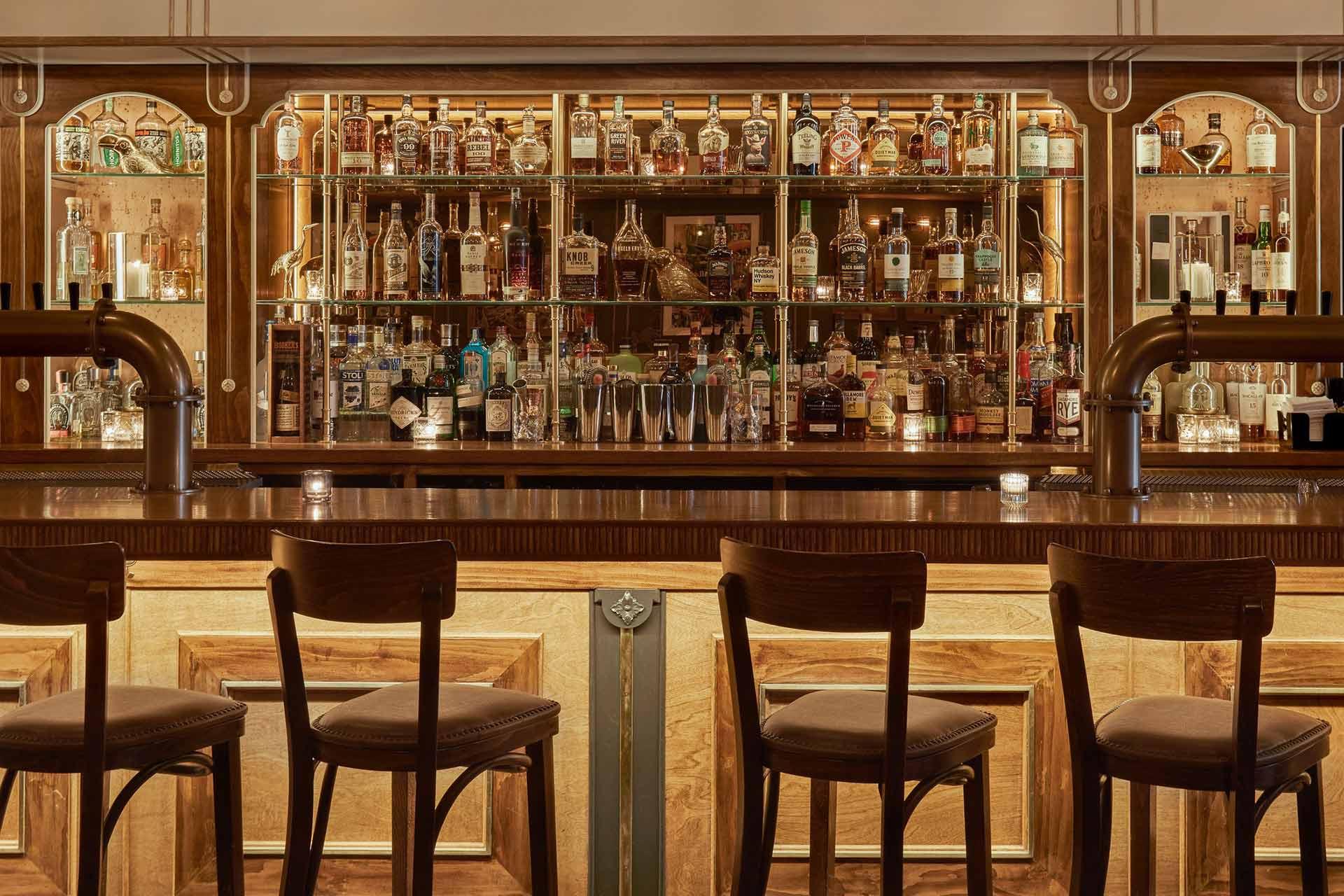 A warmly lit bar with four wooden barstools, a polished counter, and shelves behind displaying various bottles of liquor, cocktail glasses, and small lit candles.