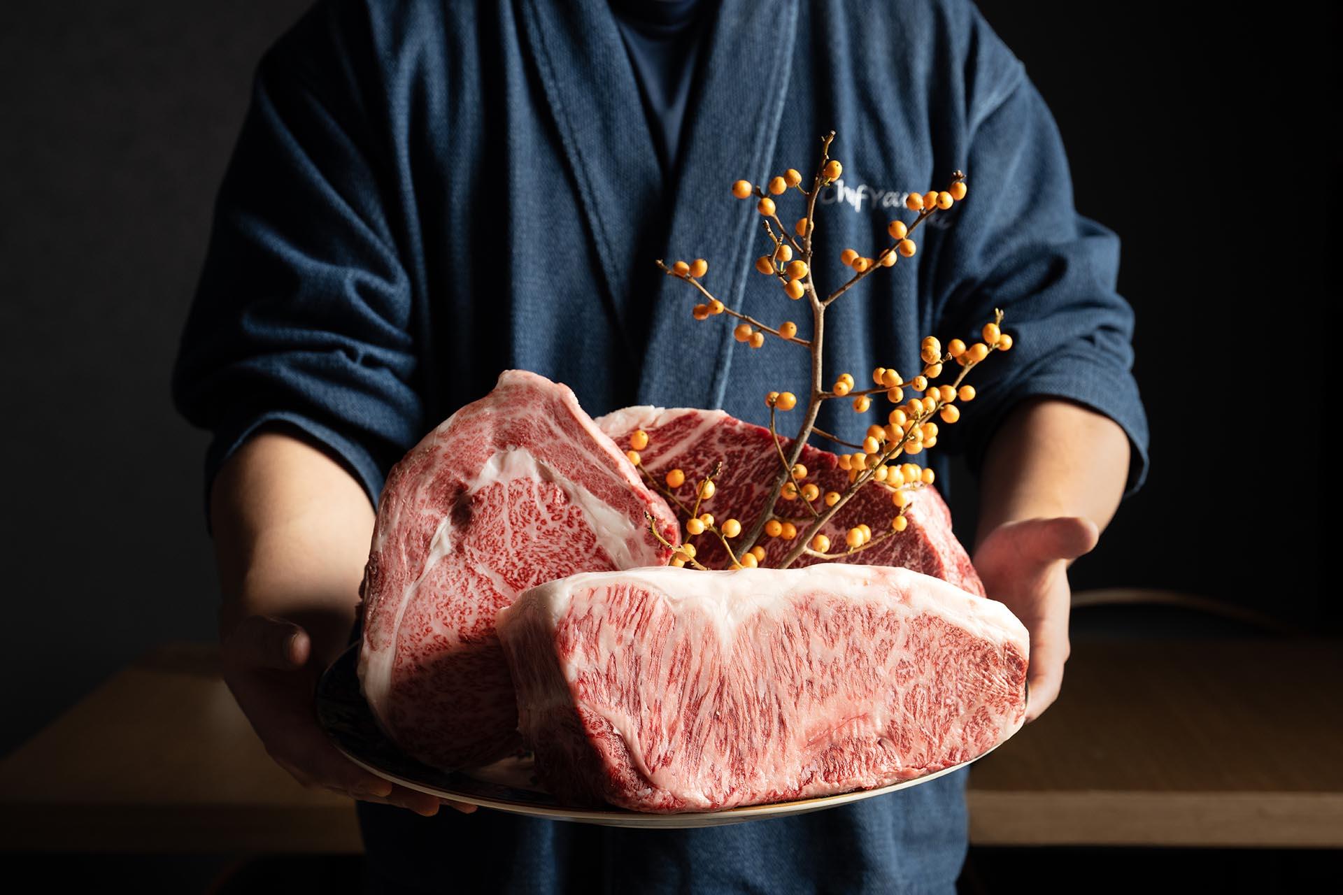 A person in a blue robe holds a large plate with three thick cuts of marbled raw beef, decorated with a sprig of orange berries.