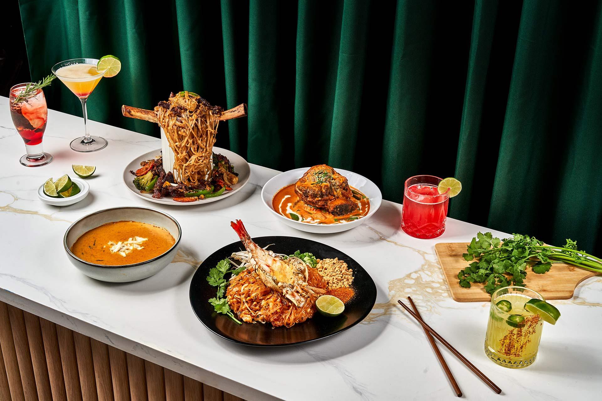 A table with colorful cocktails, a bowl of soup, a plate of noodles with shrimp, a large meat dish with noodles on top, a curry dish, fresh cilantro, lime wedges, and a green drink, all against a dark green curtain.