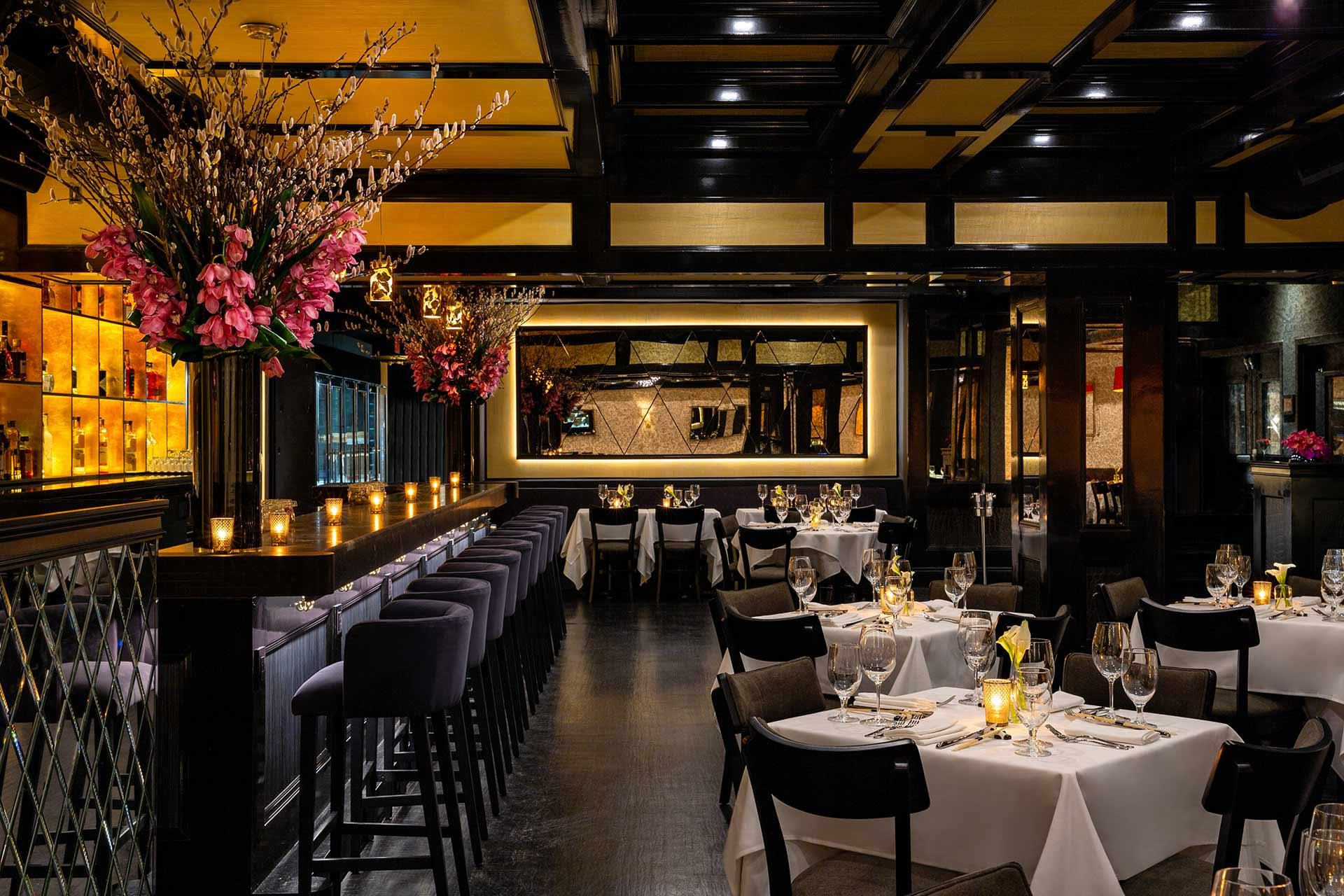 Elegant restaurant interior with dim lighting, tables set with white tablecloths and wine glasses, a long bar with high stools, floral arrangements, candles, and a large mirror on the wall creating a warm, inviting atmosphere.