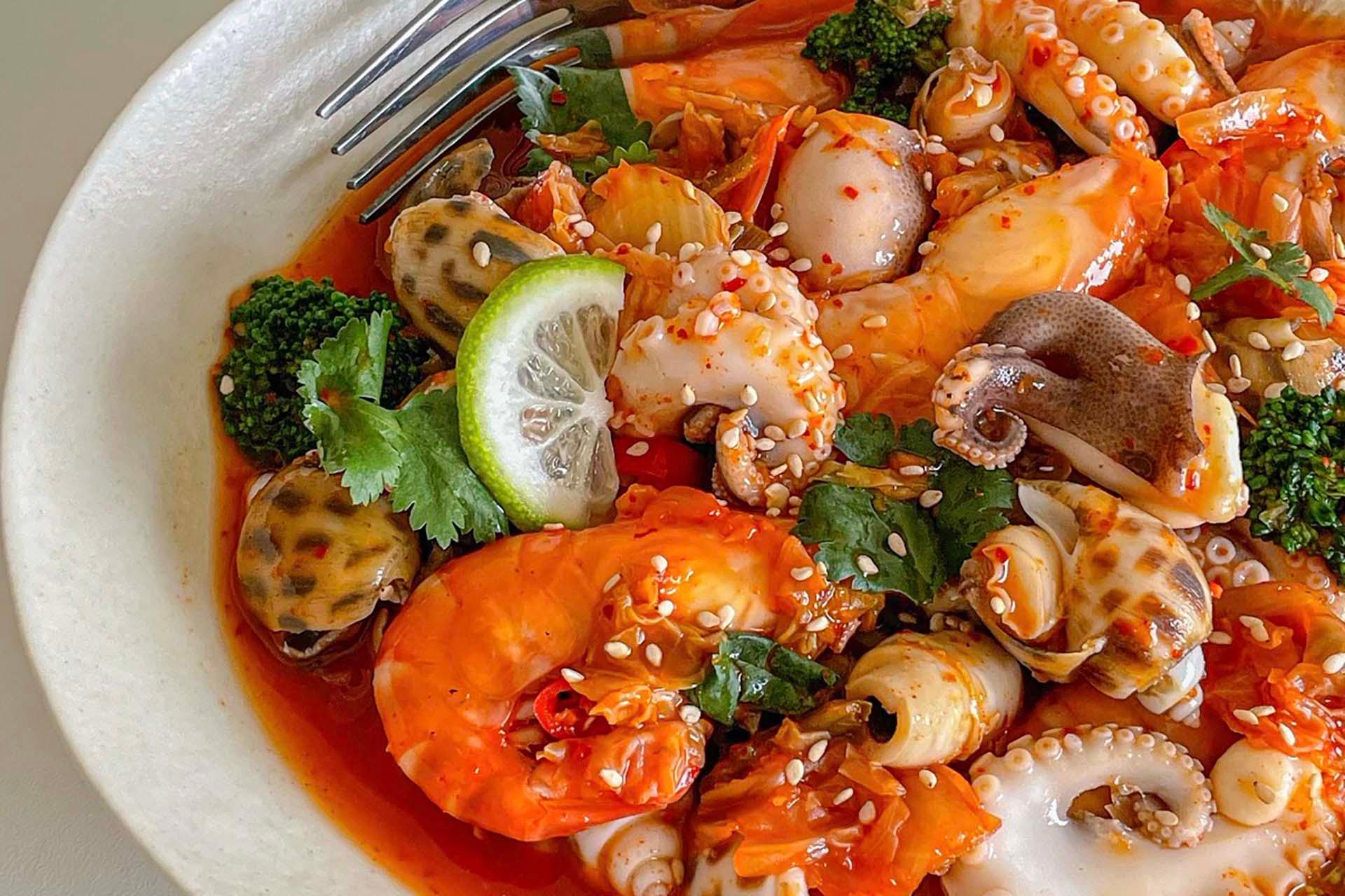 A bowl of spicy seafood stew with shrimp, octopus, clams, broccoli, cilantro, sesame seeds, and a lemon slice, with a fork on the side.