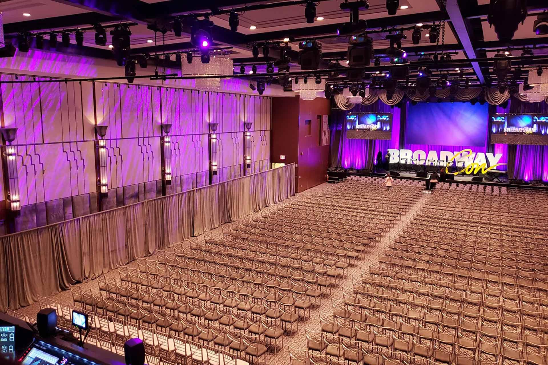 A large conference room with rows of empty chairs faces a stage with purple lighting and screens displaying "Broadway Con." The room is decorated with drapes and modern lighting fixtures.