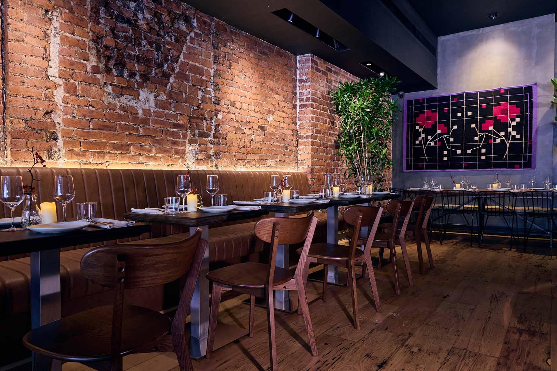 A cozy restaurant with brick walls, wooden chairs, set tables, candles, a large plant, and a colorful painting of red flowers on a black grid background. Warm lighting creates an inviting ambiance.