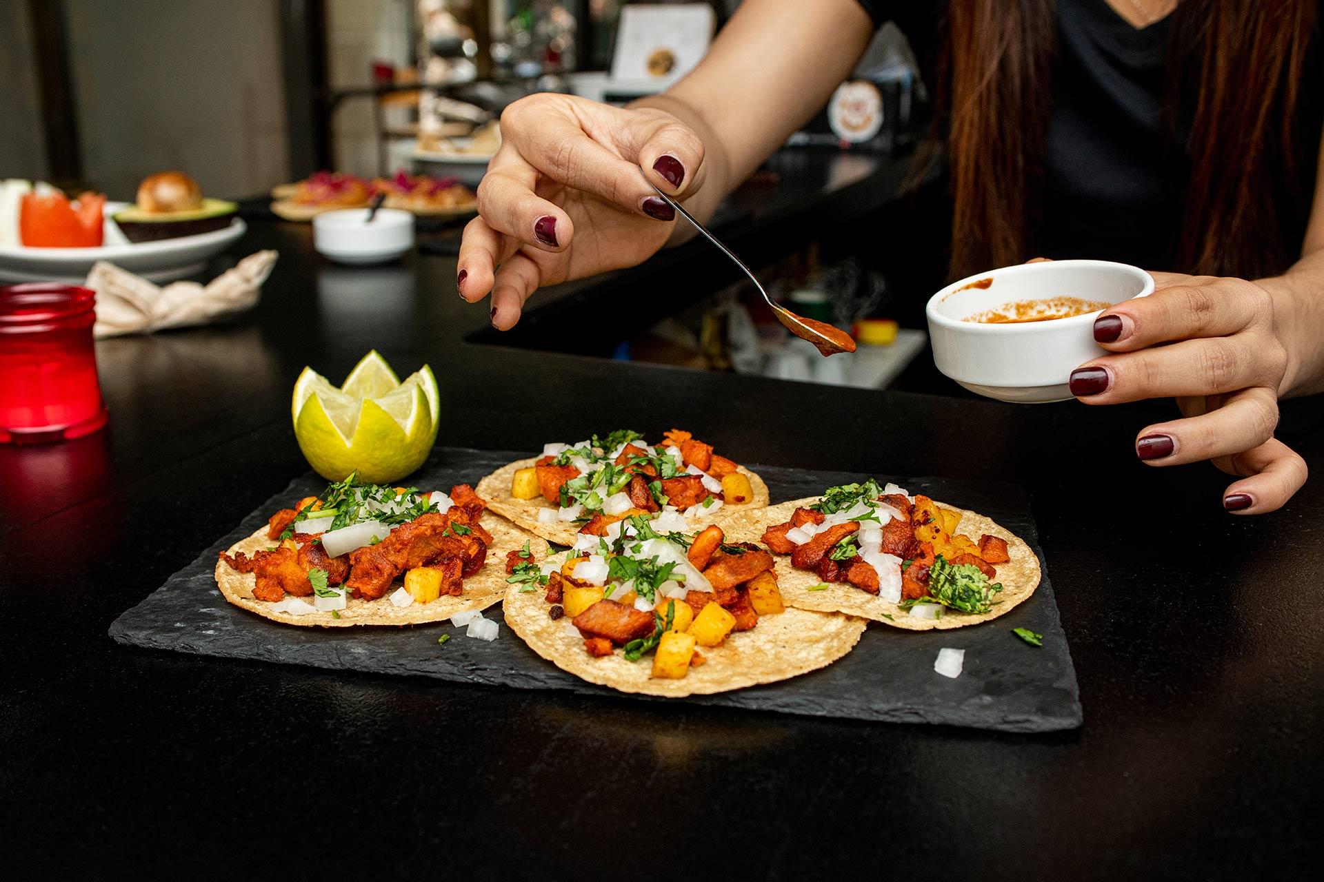 A person sprinkles seasoning onto four tacos topped with meat, onions, cilantro, and pineapple, served on a black slate plate with a decorative lime on the side.