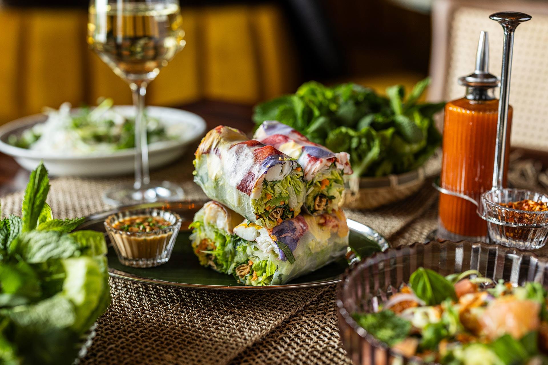 A plate of fresh spring rolls filled with vegetables and herbs, served with dipping sauce. Surrounding items include a glass of white wine, a green salad, fresh mint, and a bottle of orange sauce.