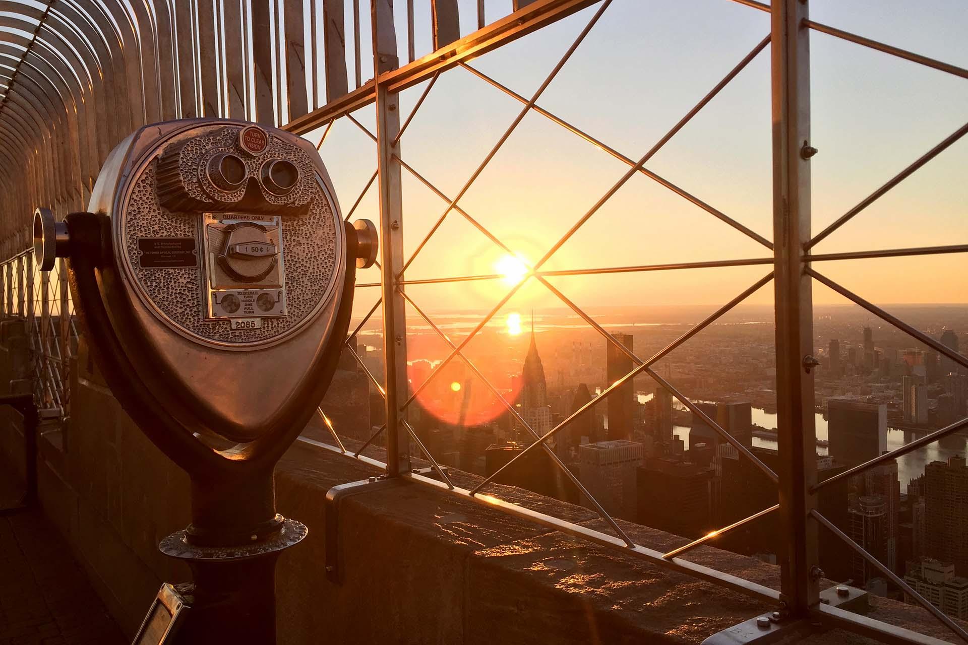A coin-operated binocular viewer on an observation deck overlooks a city skyline at sunset, with sunlight shining through a metal safety fence.