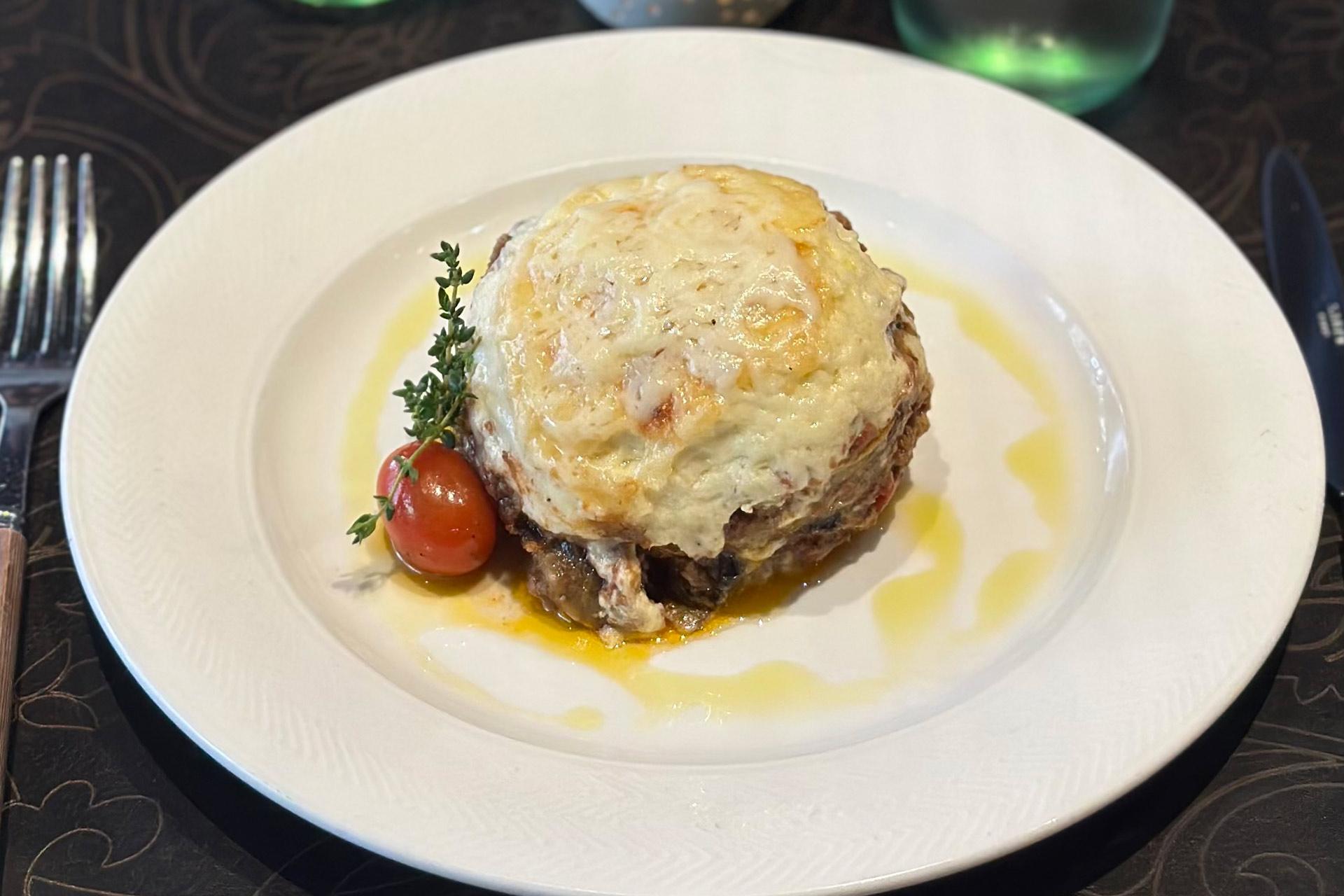 A serving of moussaka topped with melted cheese, garnished with a sprig of thyme and a cherry tomato, served on a white plate.