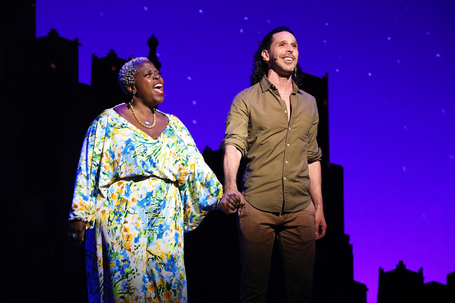 Two actors stand on stage holding hands, smiling. The woman wears a colorful floral dress and necklace, while the man wears a green shirt and brown pants. The background is purple with a city skyline and starry sky.
